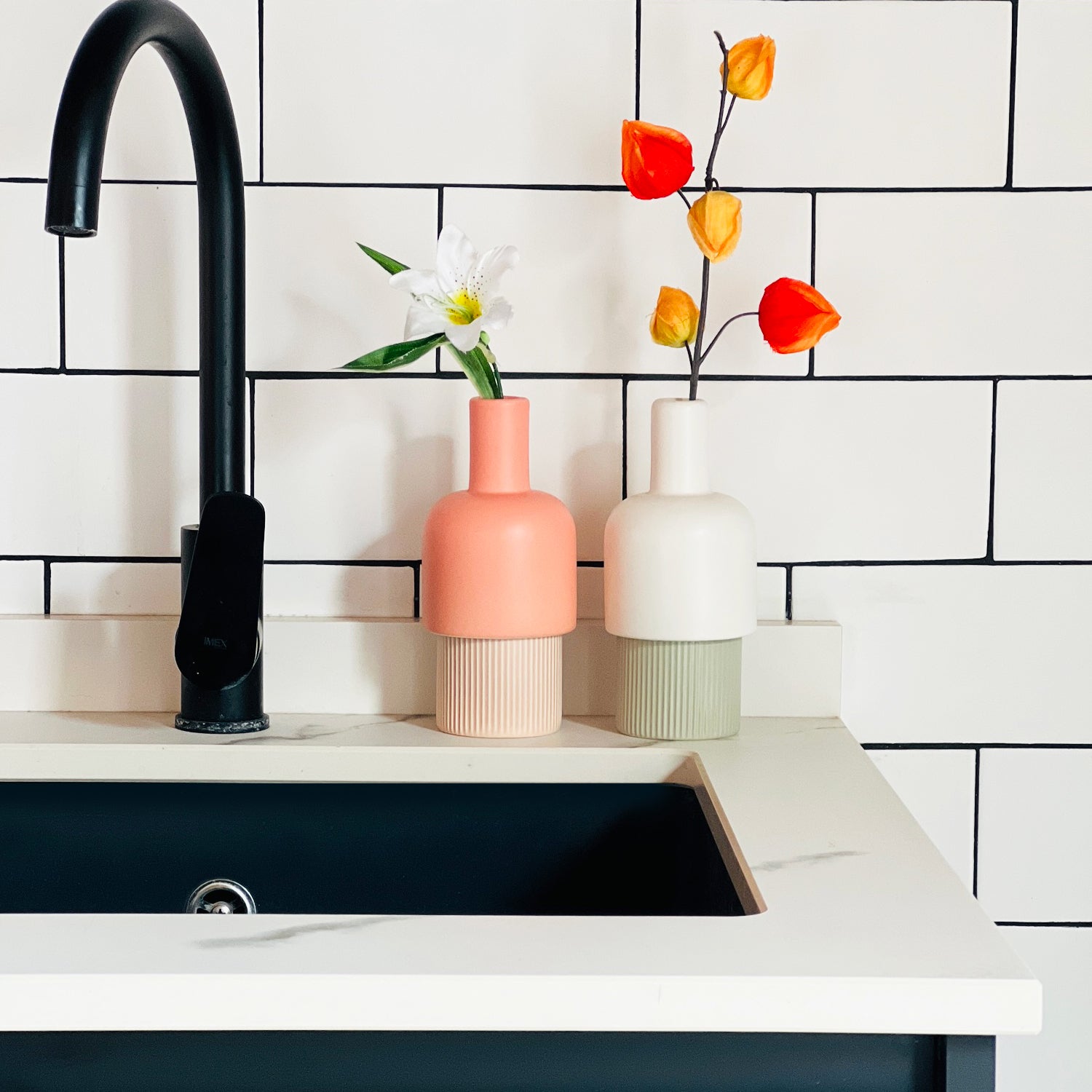 Modern kitchen sink area with black faucet, colorful vases, and flowers against a white tiled wall.