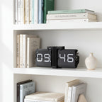 Minimalist flip clock on white shelf with stacked books, ceramic containers, and a trailing green plant, Dutch Design.
