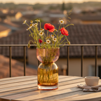 A clear glass vase with a pink upper chamber and amber lower chamber, filled with wildflowers including red poppies and white daisies, set on a wooden table outdoors with a blurred rooftop background at sunset.